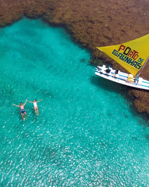 Duas pessoas boiando na água azul entre os recifes de Porto de Galinhas, ao lado de uma jangada